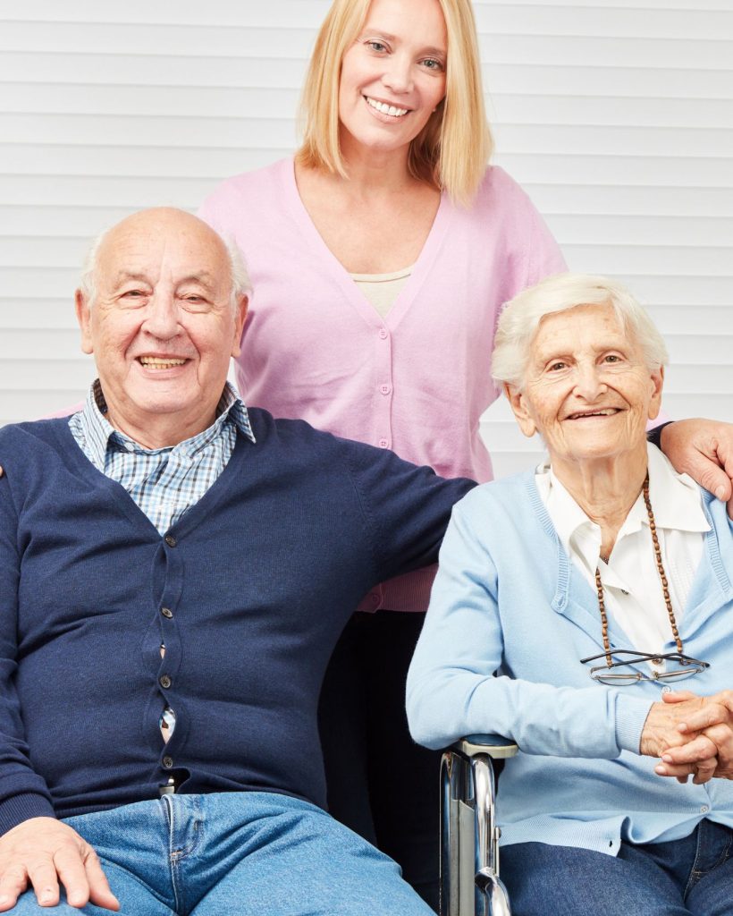 Caregiver in pink cardigan standing behind elderly man in navy sweater and elderly woman in light blue cardigan, all smiling at camera