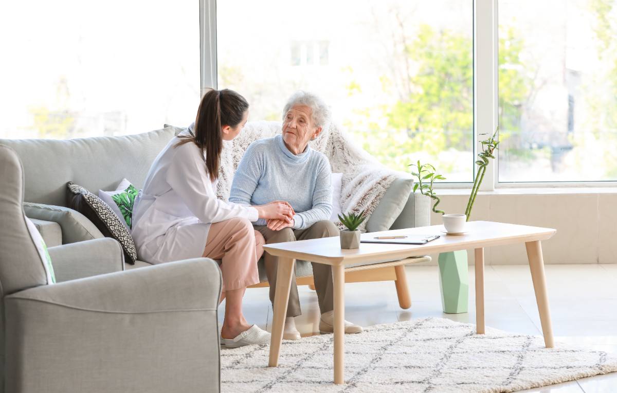 Caregiver holding hands with elderly woman on couch near bright windows