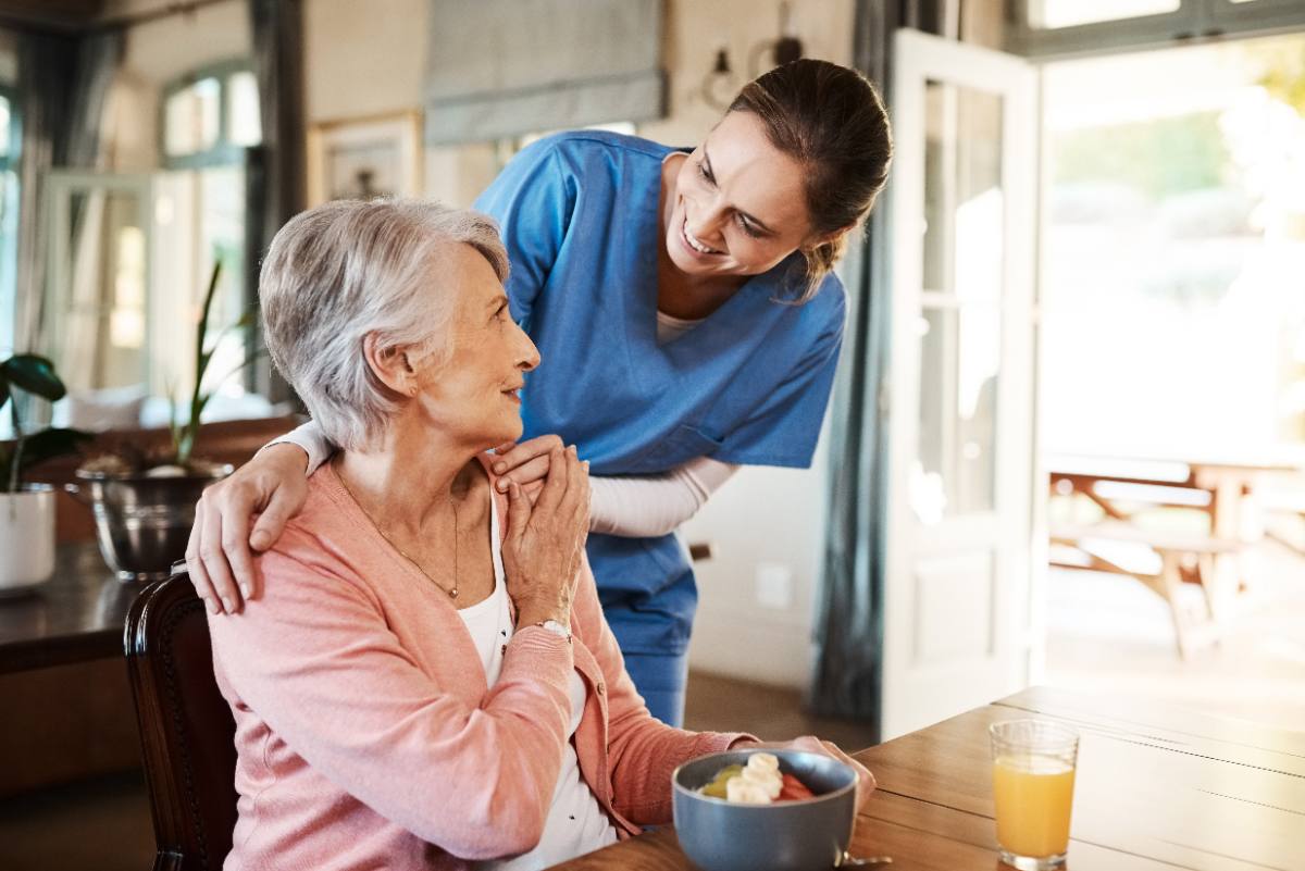 Caregiver in blue scrubs placing hand on shoulder of elderly woman in pink cardigan at dining table
